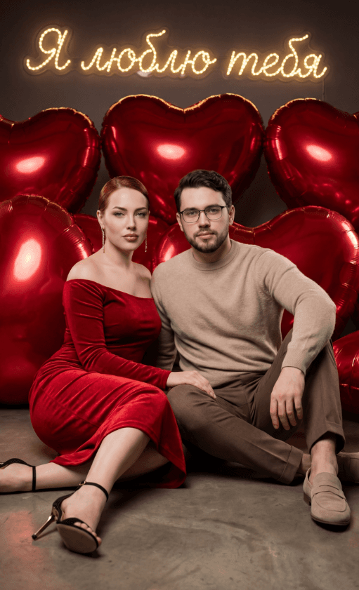 Пара сидит перед красными шарами-сердцами и неоновой надписью Я люблю тебя