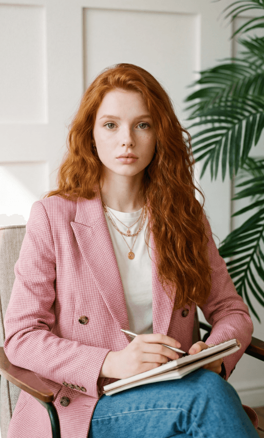 Рыжеволосая женщина в розовом пиджаке пишет в блокноте