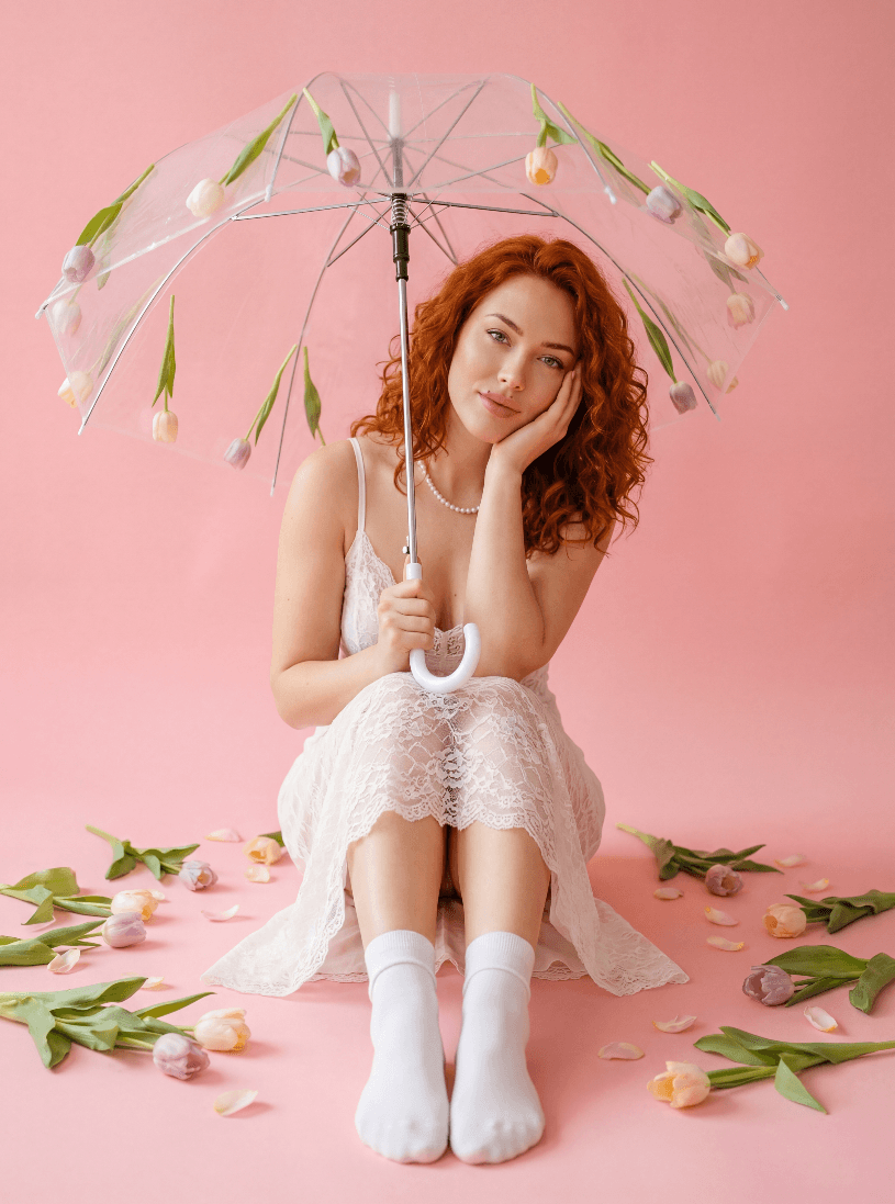 Рыжеволосая женщина в кружевном платье с прозрачным зонтом с тюльпанами на розовом фоне.