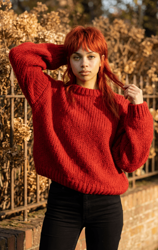 Женщина в красном вязаном свитере позирует у кованого забора осенью