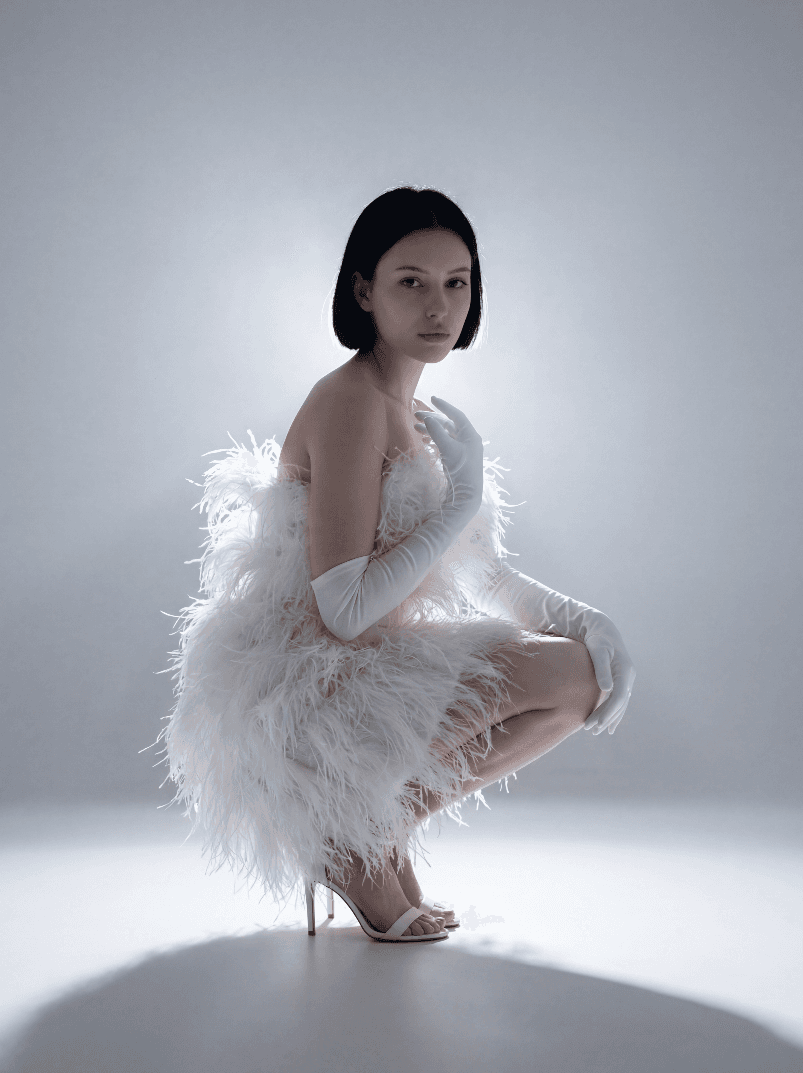 Женщина в белом перьевом платье и перчатках, присела в студии.