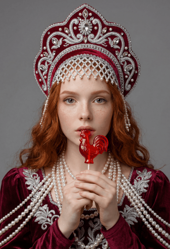 Рыжеволосая девушка в бордовом кокошнике держит леденец-петушок