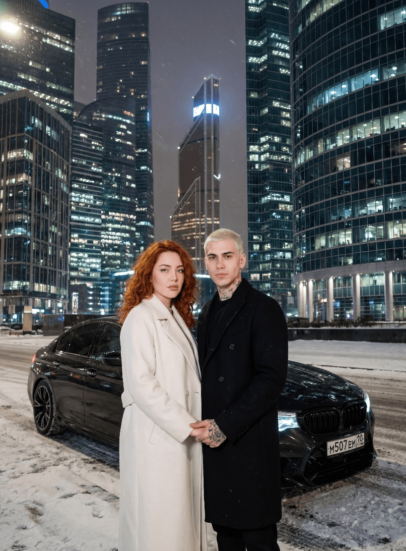 Пара у чёрного седана на заснеженной улице среди небоскрёбов ночью