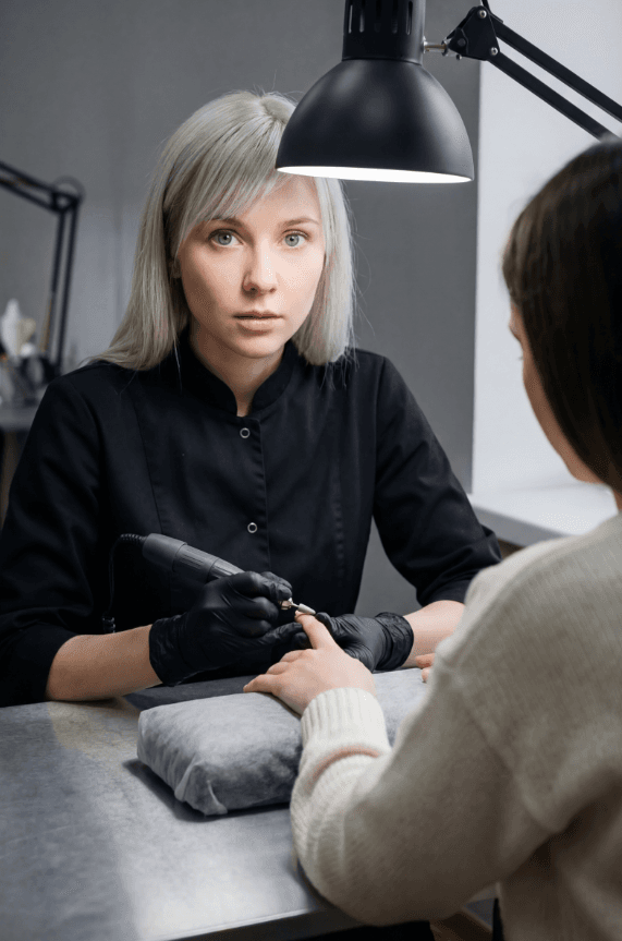 Мастер маникюра обрабатывает ногти электрическим аппаратом под лампой