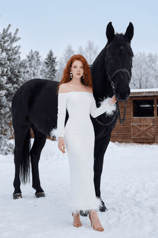 Рыжеволосая женщина в белом платье с чёрной лошадью на снегу у конюшни