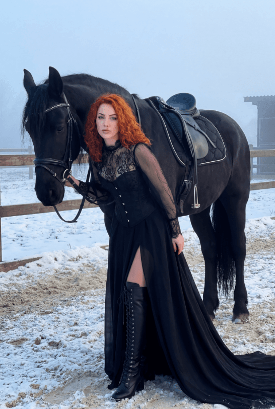 Рыжеволосая женщина в черном платье рядом с оседланной вороной лошадью на снежном паддоке