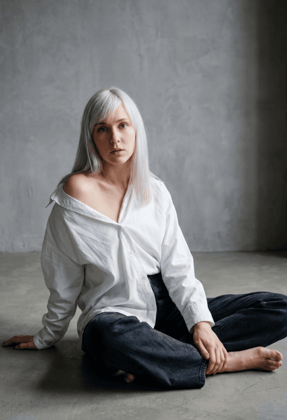 Женщина с серебристыми волосами сидит босиком на бетонном полу в белой рубашке