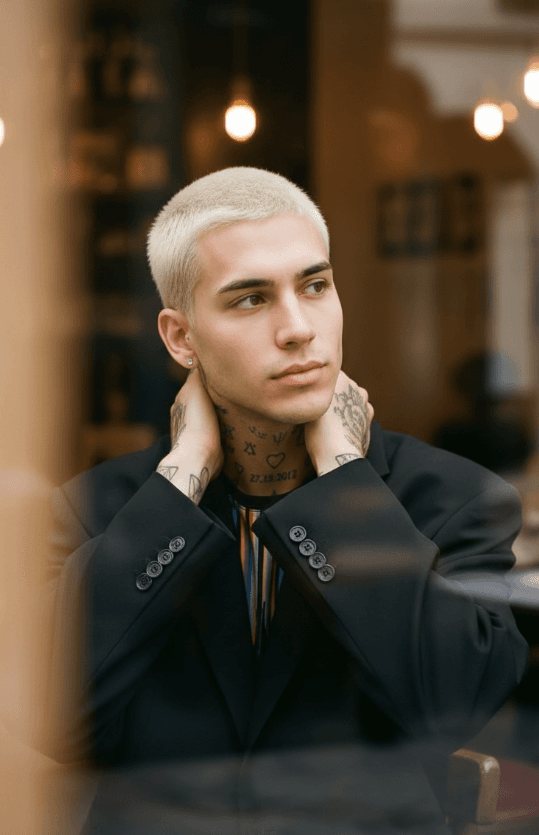 Светловолосый татуированный человек в чёрном пиджаке у окна кафе