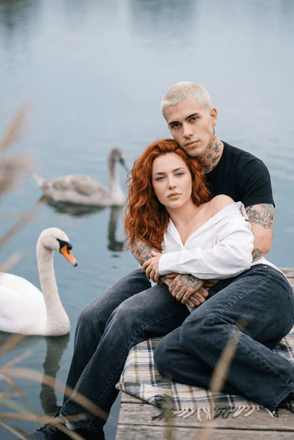 Татуированная пара обнимается на причале у озера, рядом лебеди