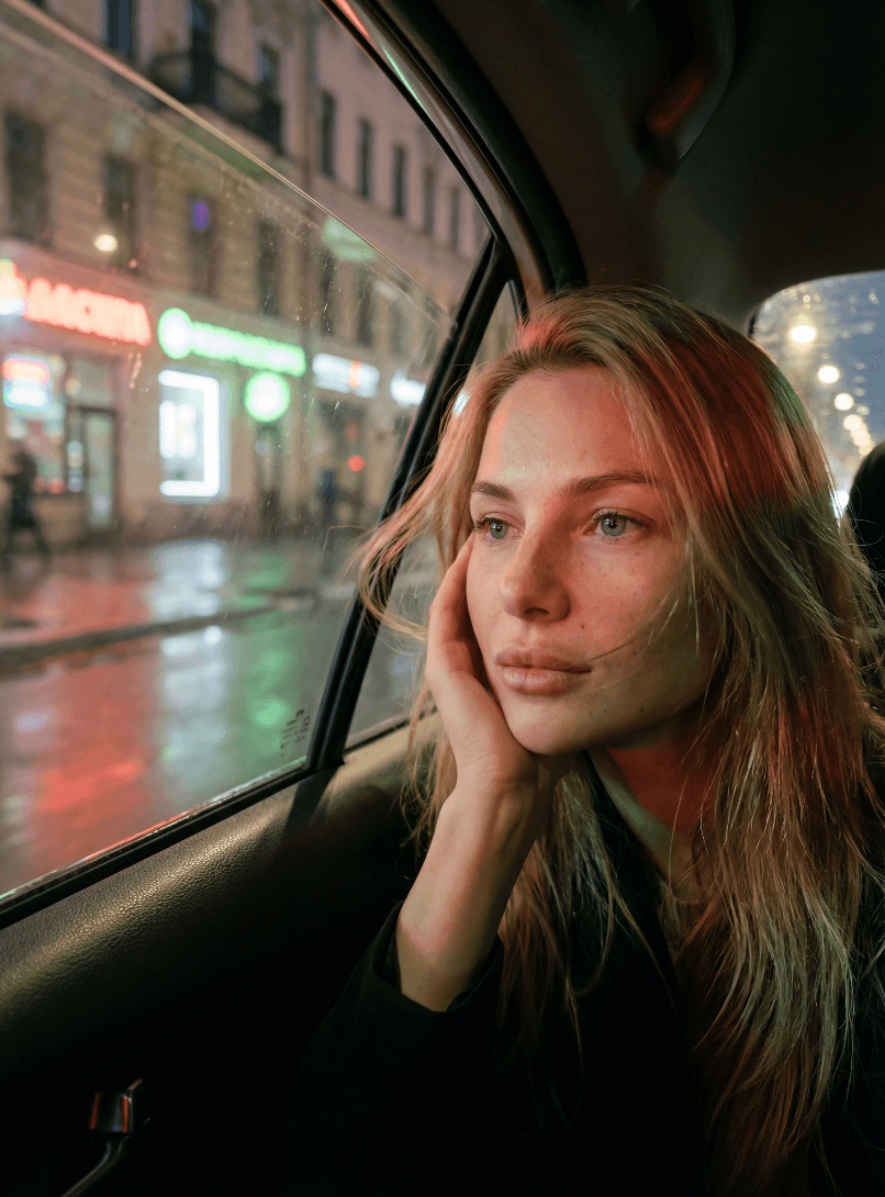 Задумчивая женщина смотрит через мокрое окно машины на огни города