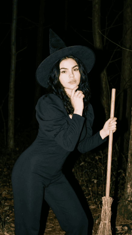 Женщина в чёрном костюме ведьмы с метлой в тёмном лесу ночью